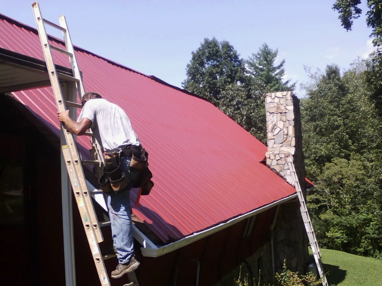 Roofer on a ladder beside a red metal roof and stone chimney — Soffit & Fascia Repair by Roofing Brothers in Abie